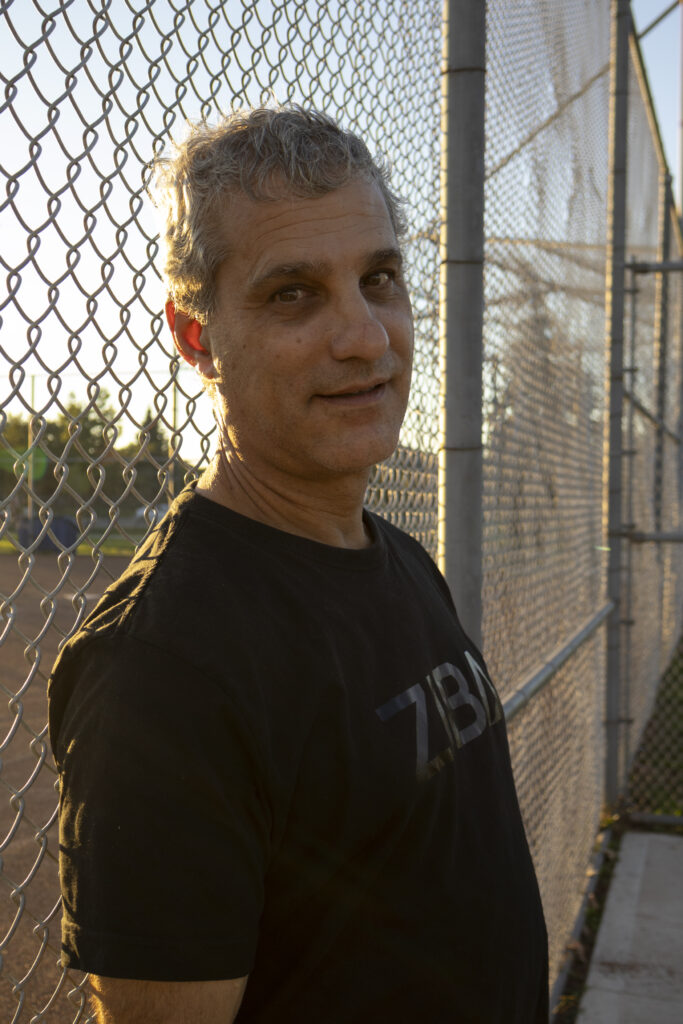 A man at golden hour leaning up against a fence of a baseball filed