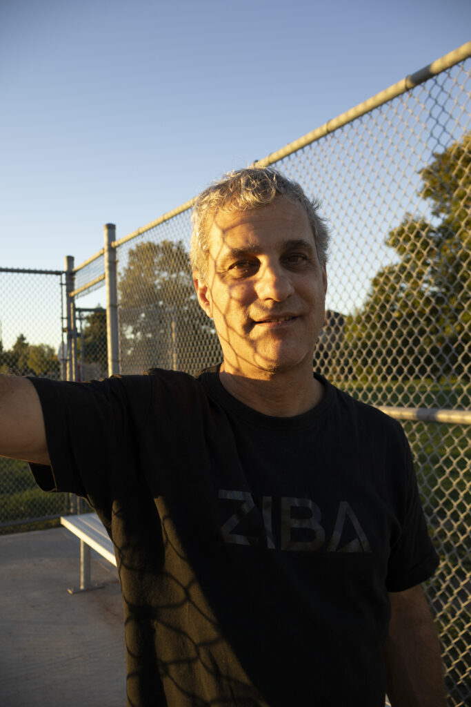 A man at golden hour leaning with arm against a fence of a baseball filed